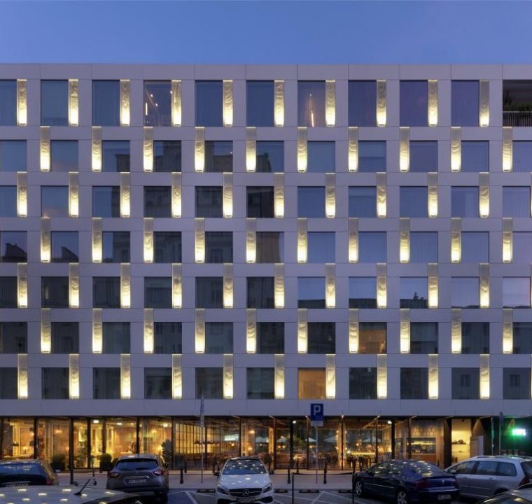 A hotel building with parking lot in front, with glass windows for each rooms and glass walls and doors on the first-level